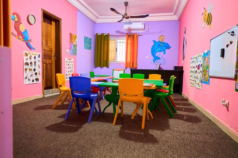Une salle de classe aux murs roses et violets : table d’activités entourée de chaises colorées, tableau blanc, affiches d’apprentissage et climatiseur.