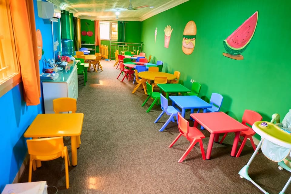 La salle de repas : petites tables et chaises colorées le long d’un mur vert décoré de fruits en relief.